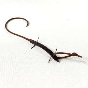 May include: A brown leather guitar strap with a curved design. The strap features a loop at one end and a series of small, thin leather pieces extending from the main body. The strap is designed to be worn over the shoulder.