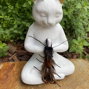 May include: A white cat figurine in a seated yoga pose with closed eyes and gold-painted ears. The cat holds a decorative feather ornament with black and brown feathers. The figurine is on a stone surface with green foliage in the background.