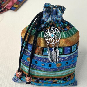 May include: A fabric drawstring pouch featuring a multi-colored geometric pattern in shades of blue, green, and gold. The bag is cinched with black cords finished with wooden beads and adorned with a silver-toned dreamcatcher charm with a turquoise center.