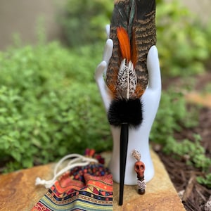 May include: A colorful feather fan with black, brown, orange, and white feathers. The fan is displayed on a white hand with a black skull charm attached to the handle. A small, colorful bag is in the foreground.