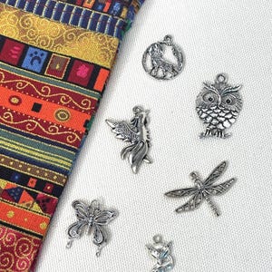 May include: A collection of six silver-toned metal charms featuring a wolf, an owl, a phoenix, a dragonfly, a butterfly, and a fox, arranged next to a piece of fabric with a multi-colored geometric and floral pattern in red, yellow, and blue.