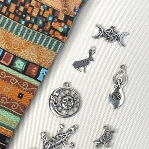 May include: A collection of six silver-toned metal charms featuring a triple moon pentacle, a rabbit, a sun and moon face, a spiral goddess figure, a lizard, and a raven, displayed next to a piece of fabric with a multi-colored geometric pattern.