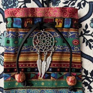 May include: A rectangular fabric pouch featuring a multi-colored horizontal striped pattern in shades of red, green, blue, and gold. A black leather drawstring closure is adorned with a silver-toned dreamcatcher charm and two wooden beads.