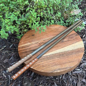 May include: Two metal chopsticks with leather-wrapped handles, one tan and one brown, are displayed on a round wooden cutting board. The handles are stitched with white thread. The background features green foliage and mulch.