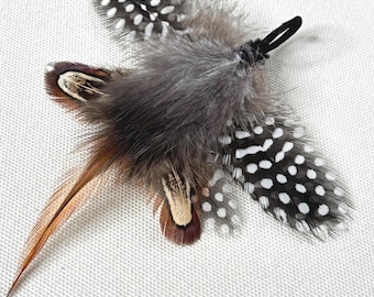 May include: A decorative feather ornament featuring a central cluster of soft, dark grey downy feathers. It includes several spotted black and white guinea fowl feathers, along with longer, pointed brown and tan striped feathers and a black loop.