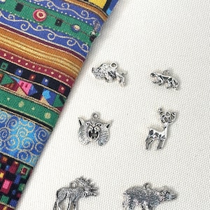 May include: Six silver-toned metal animal charms featuring a bison, a bear, a mountain lion head, a deer, a moose, and a wolf, arranged next to a corner of a multi-colored woven fabric with geometric patterns in green, blue, red, and yellow.