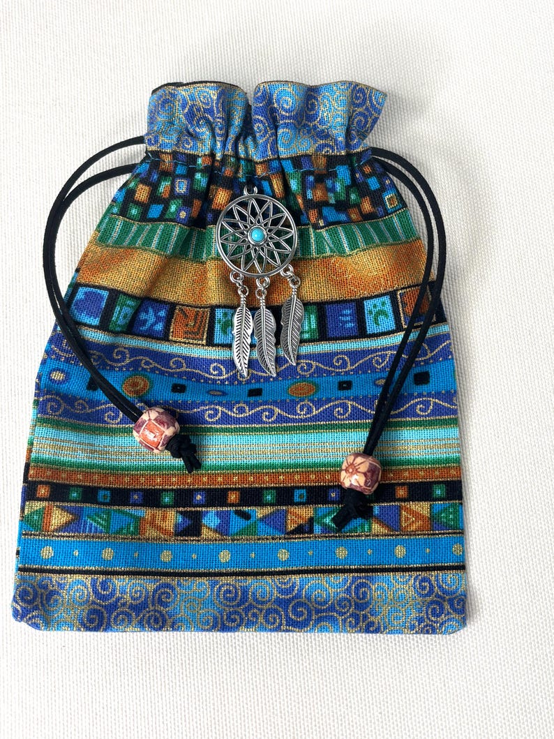 May include: A drawstring pouch featuring a multi-colored fabric with horizontal bands of geometric patterns in blue, green, gold, and black. A silver-toned dreamcatcher charm with a small turquoise bead and three feathers hangs from the top.