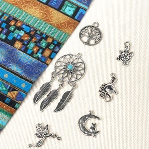 May include: A collection of silver-toned metal charms including a dreamcatcher with a turquoise center, a tree of life, a turtle, a unicorn, a fairy, and a cat sitting on a crescent moon, displayed next to a colorful fabric pouch with geometric patterns.