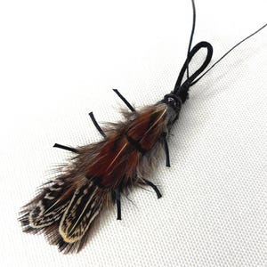 May include: A decorative feather insect with brown, black, and white feathers. The insect has a black cord and small black appendages. The feathers are arranged to resemble the body of an insect.