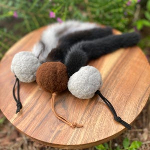 May include: A cat toy with three furry balls attached to a long, black and white tail. The balls are brown, gray, and white.