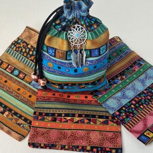 May include: A drawstring pouch made of multi-colored fabric featuring horizontal bands of geometric patterns in orange, blue, green, and gold. A silver-toned dreamcatcher charm with a small turquoise bead hangs from the black drawstring cords.
