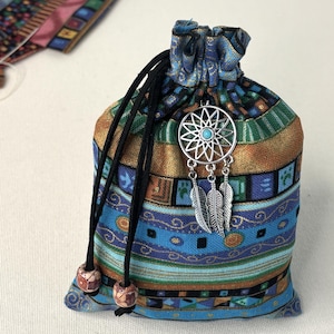 May include: A small fabric drawstring pouch featuring a multi-colored geometric pattern in shades of blue, green, and gold. The bag is cinched with black cords finished with patterned beads and adorned with a silver dreamcatcher charm.