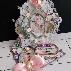 Tarjeta romántica de cumpleaños con caballete, lazo rosa, broche, muchas flores y perlas. Tarjeta personalizada: "Para mi hermosa hija".