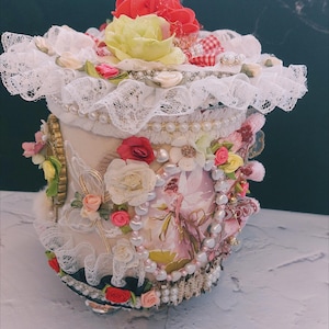 May include: A white ceramic jar decorated with lace, pearls, and fabric flowers in shades of pink, red, and white. The jar has a vintage, shabby chic style.