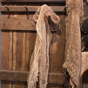 May include: A rustic scene featuring a lit candle in a dark cylindrical holder tied with twine. A tattered, beige fabric hangs from a wooden door, with another piece of fabric to the right. The candle flame glows warmly, creating a cozy, vintage aesthetic.