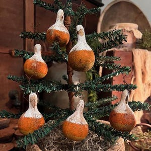 May include: A small, artificial Christmas tree adorned with six gourd-shaped ornaments. The ornaments are painted with a white top and orange body. The tree sits in a woven basket, creating a rustic, farmhouse aesthetic.