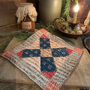 May include: A handmade, square quilt with a cross design. The quilt features a central navy blue cross with white floral accents, framed by red and tan squares. The quilt is placed on a wooden surface, with a jar of flour and a candle in the background.