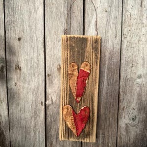 May include: A rustic wooden sign with two fabric hearts. The hearts are a combination of tan and red, attached to a weathered, rectangular wooden board. A wire loop is attached for hanging. The background is a weathered wooden wall.