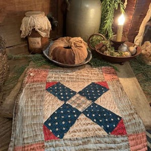 May include: A rustic, quilted placemat with a central X design in navy blue polka dot fabric, red corner accents, and a cream-colored background. The placemat is part of a vintage-themed home decor arrangement.
