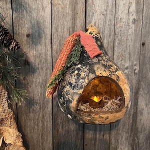 May include: A gourd-shaped decorative candle holder with a rustic, weathered appearance. The gourd is painted with black and gold tones, and has a large opening to hold a candle. It is decorated with a corn husk and greenery.