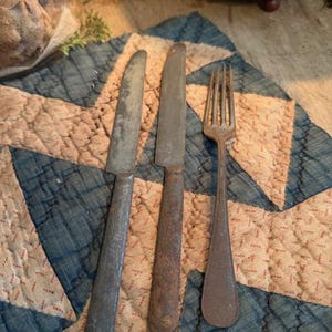 May include: Three antique metal utensils: two knives and a fork. The knives have a dull, aged appearance, with one showing a silver-gray hue and the other a rusty brown. The fork has four tines and a similar rusty finish. They rest on a blue and tan star-patterned quilt.