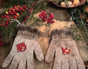 Puede incluir: Dos adornos de guantes beige con las palabras "Santa" y "Claus". Cada guante tiene un parche floral rojo y un ribete de piel sintética. Estos adornos festivos forman parte de un tema navideño.