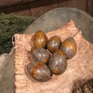 May include: Six brown and gold speckled eggs are arranged in a nest of brown fabric. The eggs are arranged in a semi-circle with the points of the eggs facing the center of the nest.