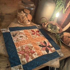May include: A square fabric quilt block featuring a central orange cross design surrounded by four star-shaped patterns in shades of red, brown, and cream. The piece is framed by a solid dark blue border and finished with visible quilted stitching.