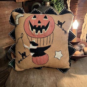 May include: A tan Halloween-themed pillow with a smiling jack-o'-lantern design. The pillow features a black and white witch riding a broom, black cats, and a crescent moon. The edges are trimmed with black sawtooth stitching.