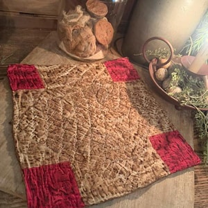 May include: A square quilted fabric mat featuring a central panel with a mottled brown and tan textured pattern. The four corners of the mat are accented with solid, vibrant red square fabric patches, all finished with visible stitching.