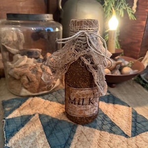 May include: A rustic, brown bottle with a label that reads "MILK". The bottle is covered in a textured material and topped with a woven net and twine. It sits on a blue and cream quilt with a geometric pattern. A glass jar and jug are in the background.
