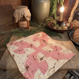 May include: A vintage quilt with a pink and white pattern, displayed on a wooden surface. The quilt features a repeating design of curved shapes. Other items in the image include a stoneware jug, a jar labeled "FLOUR", and a lit candle.