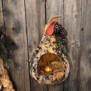 May include: A gourd-shaped candle holder with a speckled pattern in shades of brown, black, and tan. It features a cut-out opening with a small candle inside, surrounded by dried moss. Adorned with pinecones and greenery.