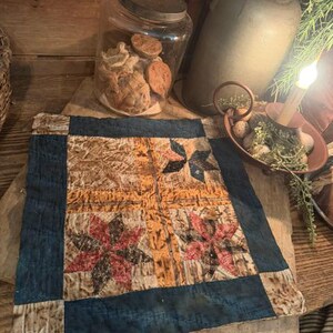 May include: A square fabric quilt block featuring a central cross design in mottled yellow, flanked by star patterns in shades of red, brown, and blue. The piece is framed by a dark blue border and displays visible hand-stitched quilting lines.