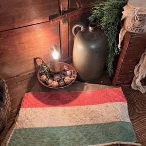 May include: A vintage flag with horizontal stripes of red, white, and green. A small candle holder with a lit candle and a decorative bowl with small, round objects are in the background. A green jug and a coffee container are also visible.