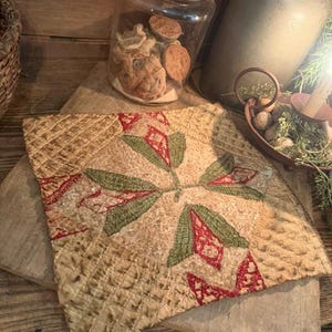 May include: A square textile piece featuring a central floral motif in muted green and red tones on a textured, beige quilted background. The fabric displays a grid-like stitched pattern throughout, with intricate needlework forming the petals and leaves.