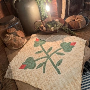 May include: A square off-white quilted fabric piece featuring a central green stem design with three green leaves and three small red flower buds. The fabric has a textured, crinkled appearance and raw, unfinished edges on all four sides.