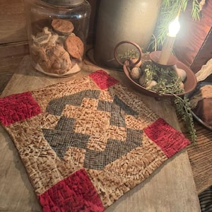 May include: A square quilted fabric mat featuring a central geometric pattern in shades of tan, brown, and dark gray, accented by solid red squares at each corner. The quilted surface displays a visible stitched texture throughout the fabric.