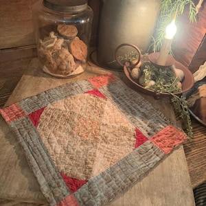 May include: A square quilted textile featuring a central beige diamond pattern surrounded by muted blue-gray borders and accented with small red corner squares. The fabric displays a visible grid of fine stitching throughout the entire surface.