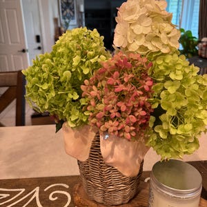 May include: A bouquet of hydrangea flowers in shades of lime green, soft pink, and cream, arranged in a textured, woven-style vase with a smooth, light-colored rim. Beside the vase sits a cylindrical candle with a silver lid and a white label.