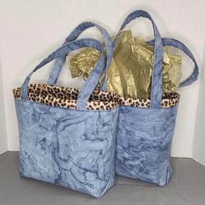 May include: Two blue denim tote bags with leopard print lining. The bags have a marbled pattern and are made of a soft, durable fabric.