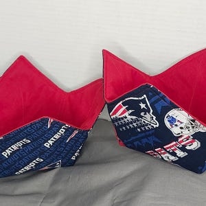 Puede incluir: Dos protectores de cuenco de tela. El interior es rojo y el exterior presenta un estampado de los Patriots en azul marino y blanco. Uno tiene la palabra "Patriots" repetida y el otro gráficos del equipo.