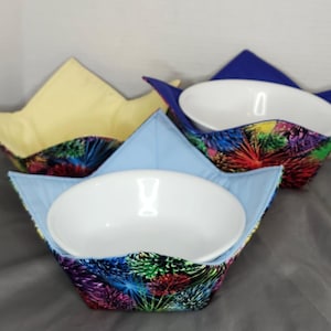 May include: Three fabric bowl cosies in different colours. The cozies are made of a colourful fabric with a firework pattern. The cozies are designed to hold bowls and keep them warm.