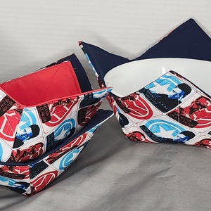 Op de afbeelding: Drie stoffen komfoorwarmers met een rood, wit en blauw patroon met een superhelden thema. De komfoorwarmers zijn gemaakt van donkerblauwe stof met een rode en witte voering.