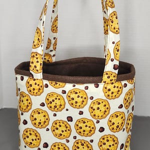May include: A white tote bag with brown handles and a chocolate chip cookie pattern. The bag has a brown lining.