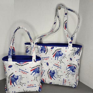 May include: Two tote bags with white fabric featuring a blue and red pattern of a buffalo breaking a table, footballs, and stars. The bags have blue lining and handles, with the text "TABLE BREAKERS" and "NO table is safe in Buffalo".