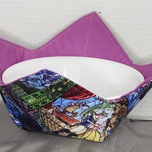 May include: A fabric bowl cosy with a purple interior and a colourful exterior featuring a stained-glass design. The cosy is designed to hold a white bowl. The exterior design includes images of characters.