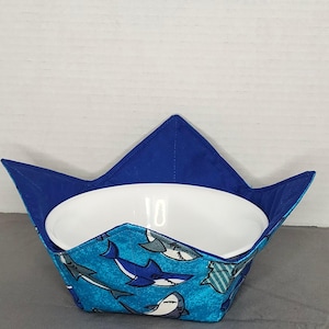 May include: A blue fabric bowl cozy with a shark pattern, holding a white ceramic bowl. The cozy has a blue exterior and a shark-themed interior. The bowl is centered within the cozy, which has a pointed, crown-like shape.