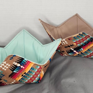 May include: Two fabric bowl cozies with a Southwestern pattern. The exterior features a brown background with geometric shapes in red, turquoise, yellow, and black. One cozy has a turquoise interior, the other a tan interior.