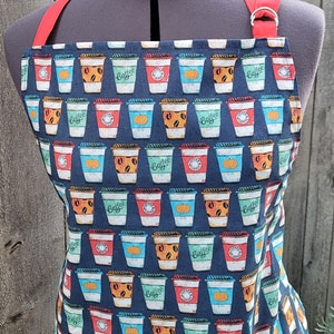Puede incluir: Un delantal azul y negro con un patrón repetido de tazas de café. Las tazas son de varios colores y tienen la palabra "Coffee" escrita en ellas. El delantal tiene una correa roja y un bolsillo blanco.